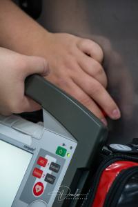 Nahaufnahme eines Defibrillators mit Bedienknöpfen und grüner Leuchtanzeige, daneben liegt eine Hand.