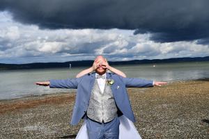Ein Bräutigam, in einem hellblauen Anzug, steht am Kiesstrand mit ausgestreckten Armen, während eine Braut hinter ihm steht und seine Augen bedeckt; dunkle Wolken im Hintergrund.
