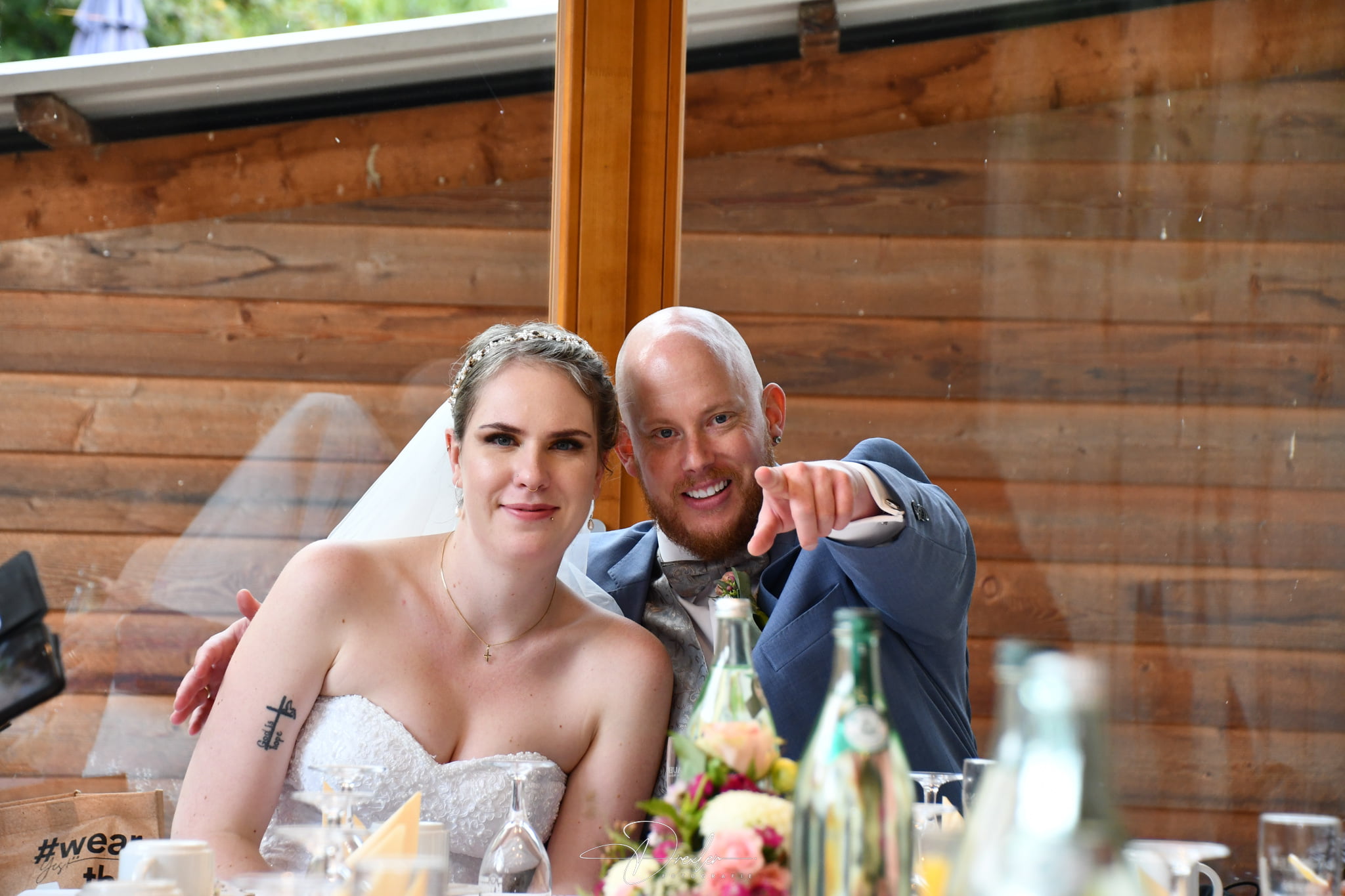 Event Fotografie 2 Ein Brautpaar sitzt an einem Tisch, der Bräutigam zeigt in die Kamera.