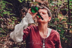 Frau in mittelalterlicher Kleidung trinkt aus einer grünen Flasche im Wald.