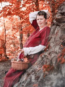 Eine Person in mittelalterlicher Kleidung lehnt an einem Felsen im Wald mit Herbstlaub und hält einen Korb.