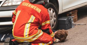 Feuerwehrmann kniet neben einer Person, die unter einem Auto liegt.