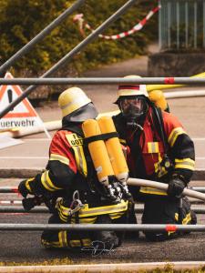 Zwei Feuerwehrleute in Schutzkleidung und Helmen, kniend mit Atemschutzgeräten und einem Feuerwehrschlauch.