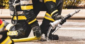 Ein Feuerwehrmann in Schutzkleidung kniet auf dem Boden und hält einen Feuerwehrschlauch, aus dem Wasser fließt.