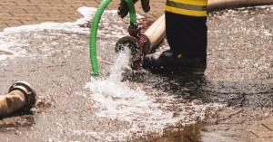 Ein Feuerwehrmann in schwarzer Uniform mit gelben Streifen hält einen Wasserschlauch, aus dem Wasser auf den Boden fließt.