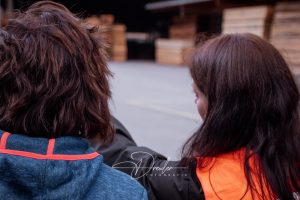 Zwei Personen von hinten, eine trägt eine blaue Jacke, die andere eine orange Warnweste, im Hintergrund Holzstapel.
