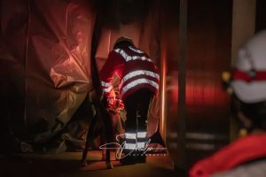Rettungskraft in reflektierender Warnkleidung und Helm mit einem Hund in einem dunklen Raum mit orangerotem Licht.