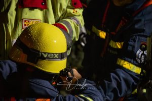 Ein Feuerwehrmann mit gelbem Helm steht im Regen, umgeben von anderen Einsatzkräften.