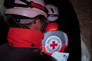 Hinteransicht von zwei Personen in Schutzkleidung mit Helmen, eine trägt eine Jacke mit dem Logo des Deutschen Roten Kreuzes.