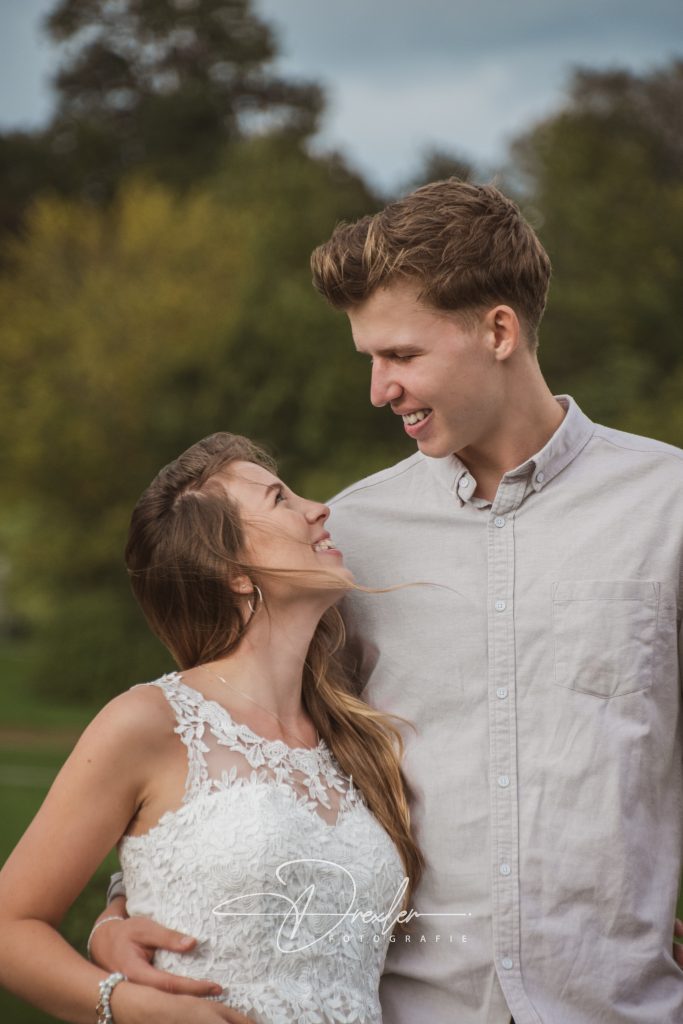 Ein Paar steht in einem Garten, die Frau trägt ein weißes Kleid mit Spitzenmuster, der Mann trägt ein hellgraues Hemd.