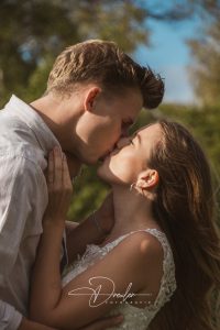 Ein junges Paar küsst sich im Freien, mit einem Mann in einem weißen Hemd und einer Frau in einem Spitzenoberteil, umgeben von grünem Hintergrund.