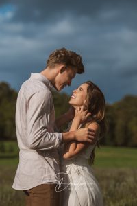 Ein Paar steht auf einem Feld bei bewölktem Himmel; der Mann trägt ein helles Hemd und die Frau ein weißes Kleid, sie schauen sich lächelnd an.