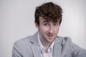 Ein Mann mit lockigem Haar trägt ein hellgraues Jackett und ein weißes Hemd vor einem neutralen Hintergrund.