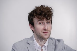 Ein Mann mit lockigem Haar trägt ein hellgraues Jackett und hat einen dezenten Gesichtsausdruck vor einem neutralen Hintergrund.