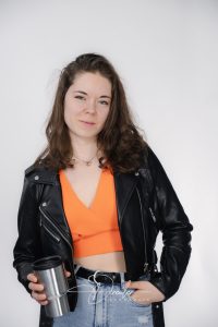 Junge Frau in schwarzer Lederjacke und orangefarbenem Top hält einen silbernen Thermobecher in der Hand.