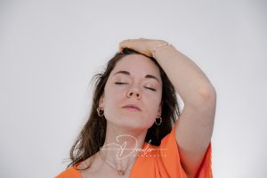 Frau in einem orangefarbenen Oberteil mit geschlossenen Augen hält eine Hand auf ihrem Haar.