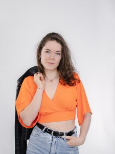 Eine Frau mit langen Haaren trägt ein orangefarbenes, bauchfreies Top und eine blaue Jeans mit Gürtel. Sie hält eine schwarze Jacke über ihrer Schulter und schaut direkt in die Kamera.