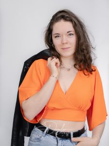 Frau in einem orangefarbenen Oberteil mit kurzen Ärmeln, Jeans und einer schwarzen Jacke über der Schulter.
