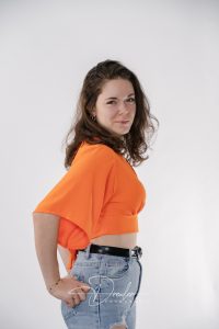 Frau in einem orangefarbenen Oberteil und blauen Jeans posiert vor einem neutralen Hintergrund.