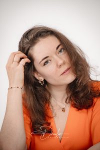 Porträt einer Frau mit braunem Haar und orangefarbenem Oberteil, die ihre Hand in ihrem Haar hat und freundlich in die Kamera schaut.