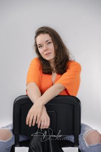 Junge Frau in einem orangefarbenen Oberteil sitzt verkehrt herum auf einem Stuhl, ihre Arme über die Stuhllehne gelegt, mit einem neutralen Gesichtsausdruck.