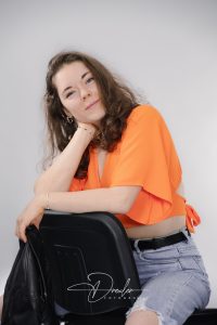 Eine Frau mit braunen Haaren trägt ein leuchtend oranges Oberteil und sitzt lässig auf einem Stuhl, lächelt in die Kamera.
