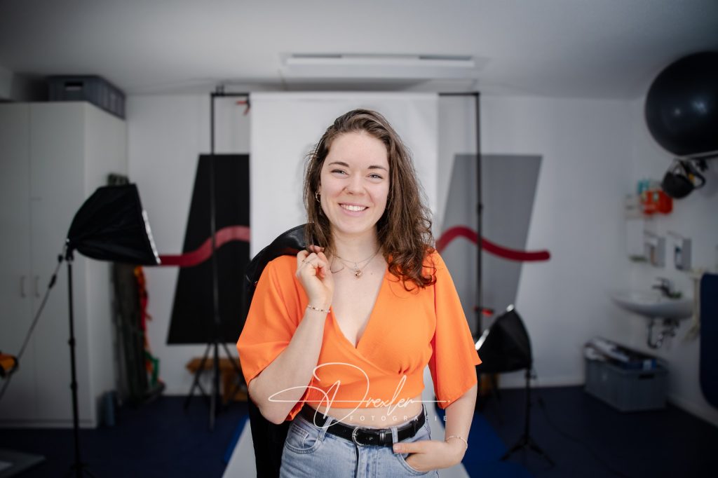 Frau mit lockigem Haar und orangefarbenem Oberteil lächelt in einem Fotostudio mit Beleuchtungsequipment im Hintergrund.