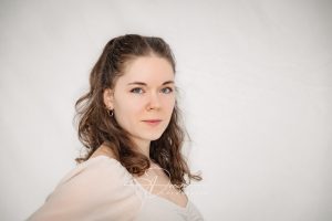 Porträt einer Frau mit lockigem Haar und Nasenpiercing, die in die Kamera schaut, vor einem hellen Hintergrund.