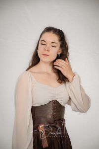 Eine Frau in einem mittelalterlich inspirierten Outfit mit geschlossenem Oberteil und Lederkorsett hält nachdenklich eine Hand an ihr Haar.