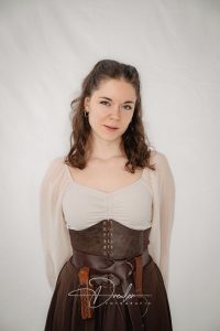 Frau mit lockigem, braunem Haar trägt ein mittelalterliches Kostüm mit Bluse und Mieder vor hellem Hintergrund.