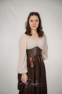 Frau in mittelalterlich inspiriertem Kostüm mit cremeweißem Oberteil und lederner Korsage, vor neutralem Hintergrund.