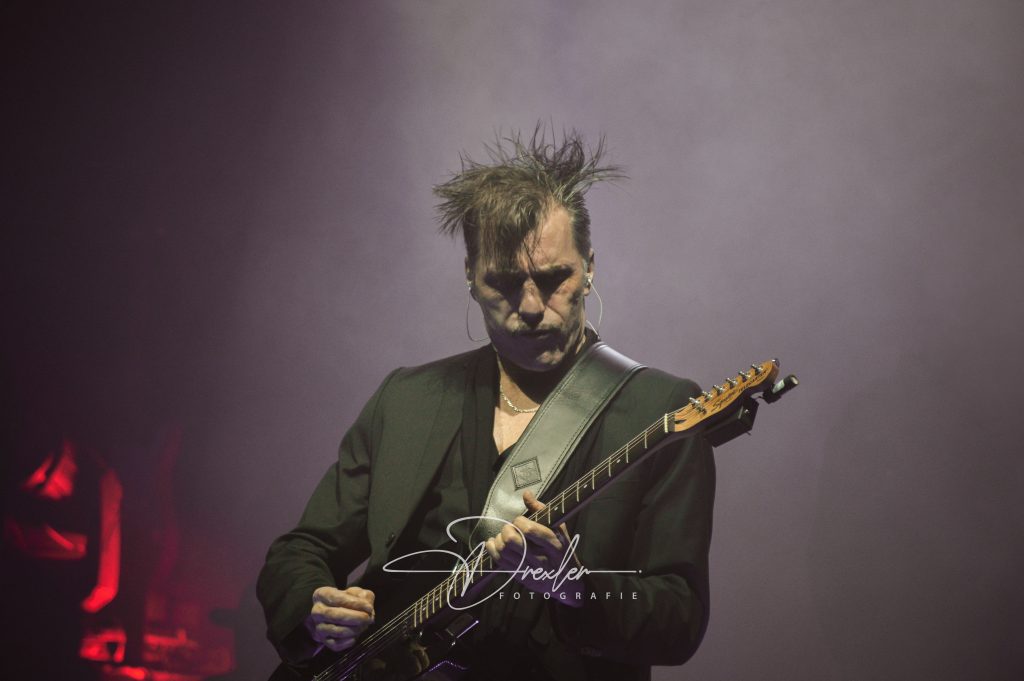 Ein Musiker mit zerzaustem Haar trägt einen schwarzen Anzug und spielt leidenschaftlich eine elektrische Gitarre auf einer Bühne mit nebliger Beleuchtung.