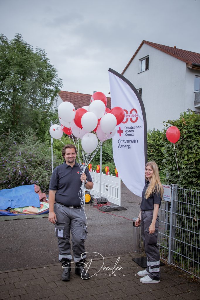 Zwei Personen stehen bei einer Veranstaltung, der Mann hält eine Gruppe von roten und weißen Ballons, im Hintergrund ist ein Banner des Deutschen Roten Kreuzes und einige dekorative Elemente sichtbar, die typisch für ein Fest sind.