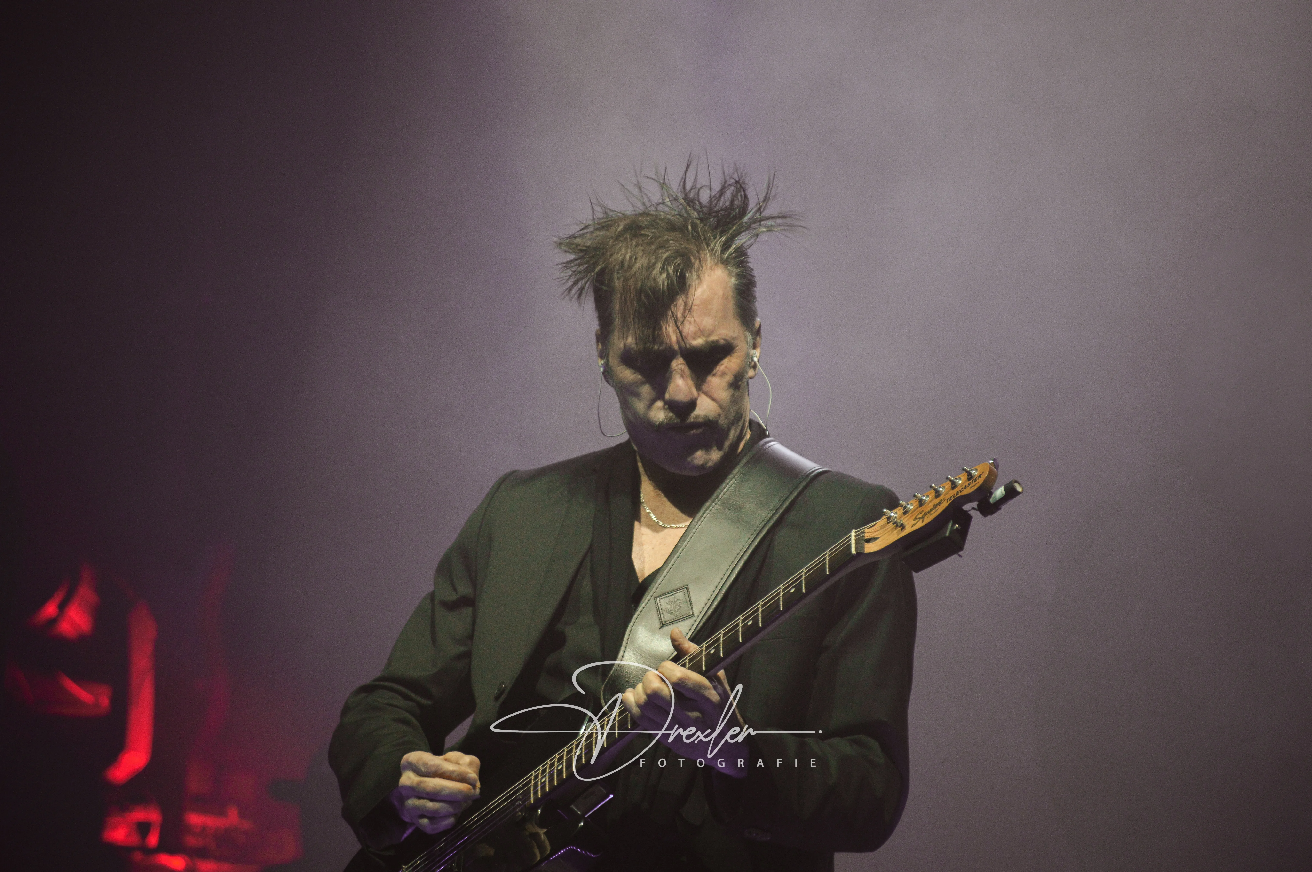 Ein Musiker mit zerzaustem Haar trägt einen schwarzen Anzug und spielt leidenschaftlich eine elektrische Gitarre auf einer Bühne mit nebliger Beleuchtung.