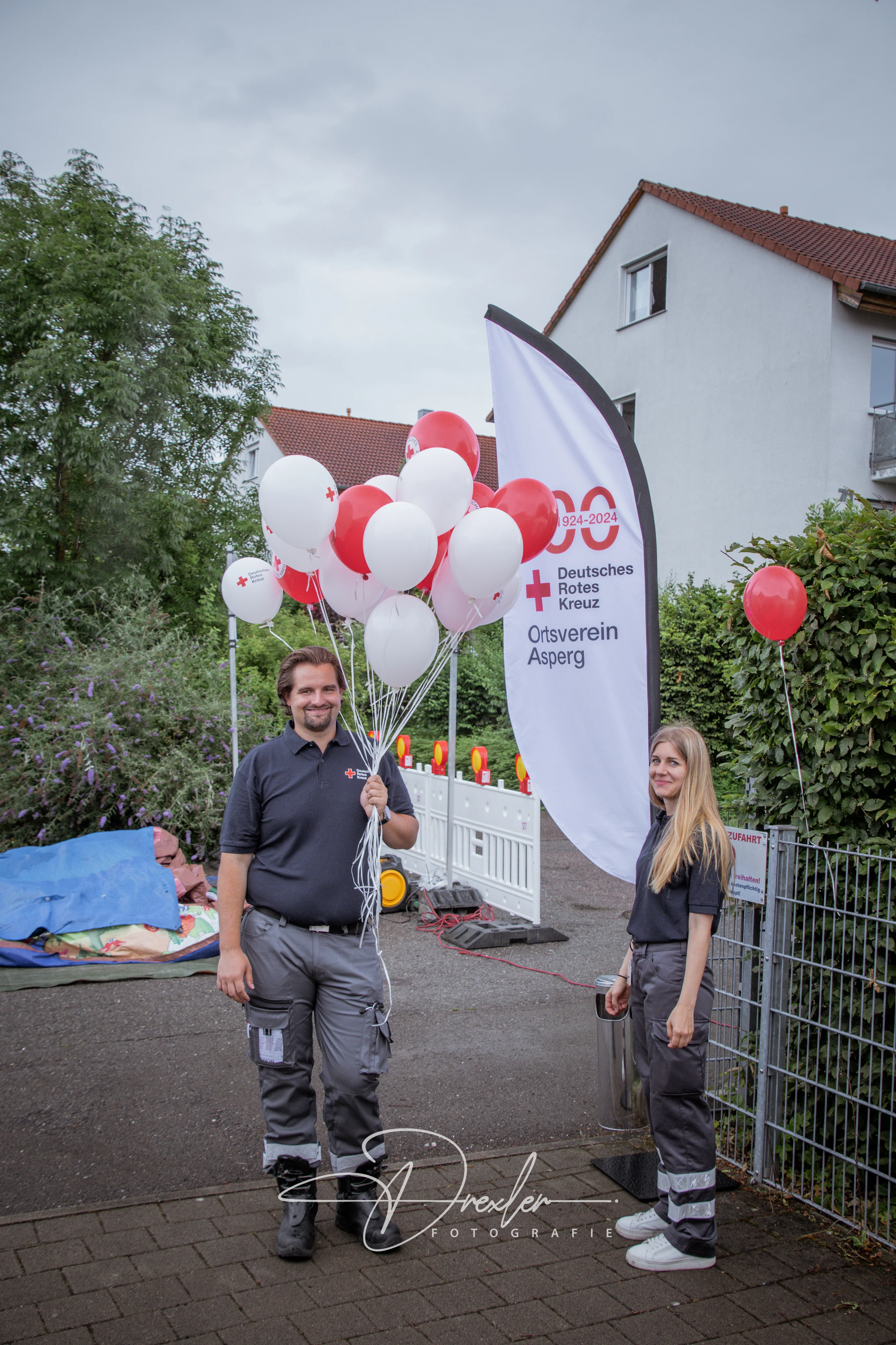 Zwei Personen stehen bei einer Veranstaltung, der Mann hält eine Gruppe von roten und weißen Ballons, im Hintergrund ist ein Banner des Deutschen Roten Kreuzes und einige dekorative Elemente sichtbar, die typisch für ein Fest sind.